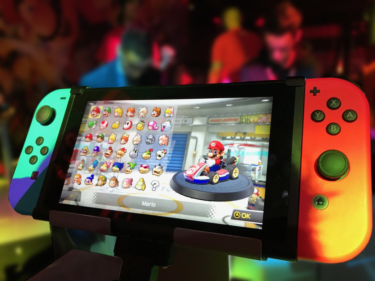 Offerings Close-up of a Nintendo Switch showing Mario Kart selection screen, gaming atmosphere in the background.